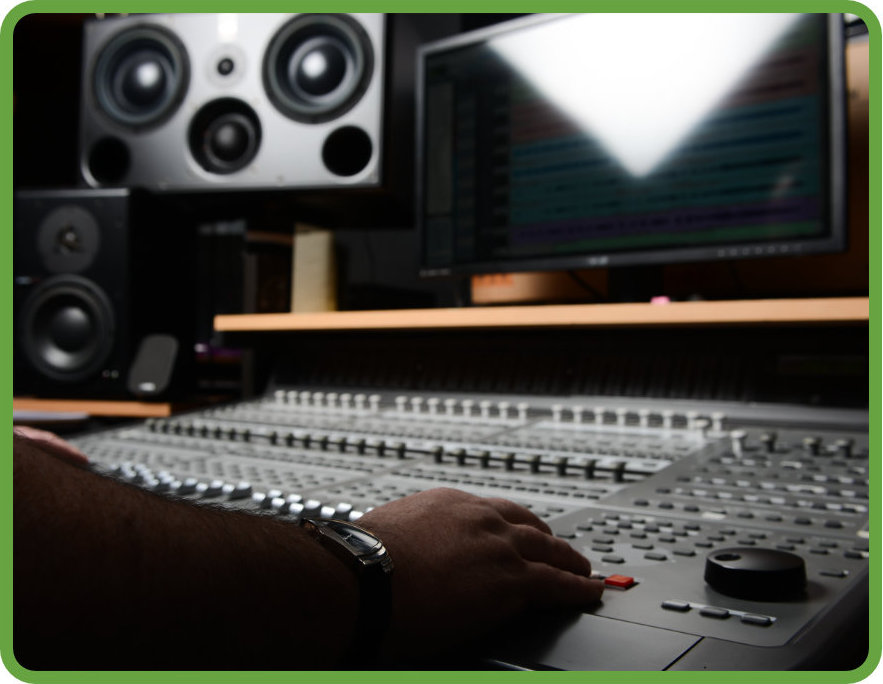 An engineer sits at a mixing desk and slides a fader forward. An audio interface is displayed on a monitor in front of him.